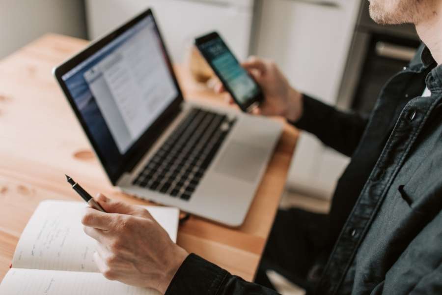 Un homme consultant son smartphone et prenant des notes devant son laptop ouvert, dans le processus d'acheter un laptop en ligne.