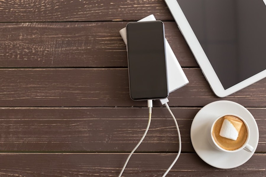 Smartphone noir branché à une batterie externe blanche sur une table en bois sombre à côté d'un café.