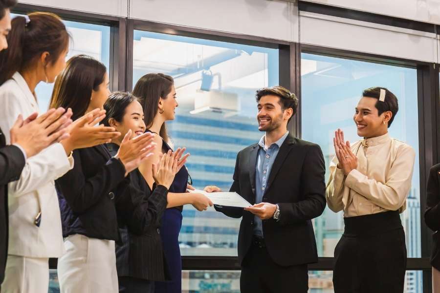 Un professionnel en costume reçoit un diplôme sous les applaudissements de ses collègues de bureau.